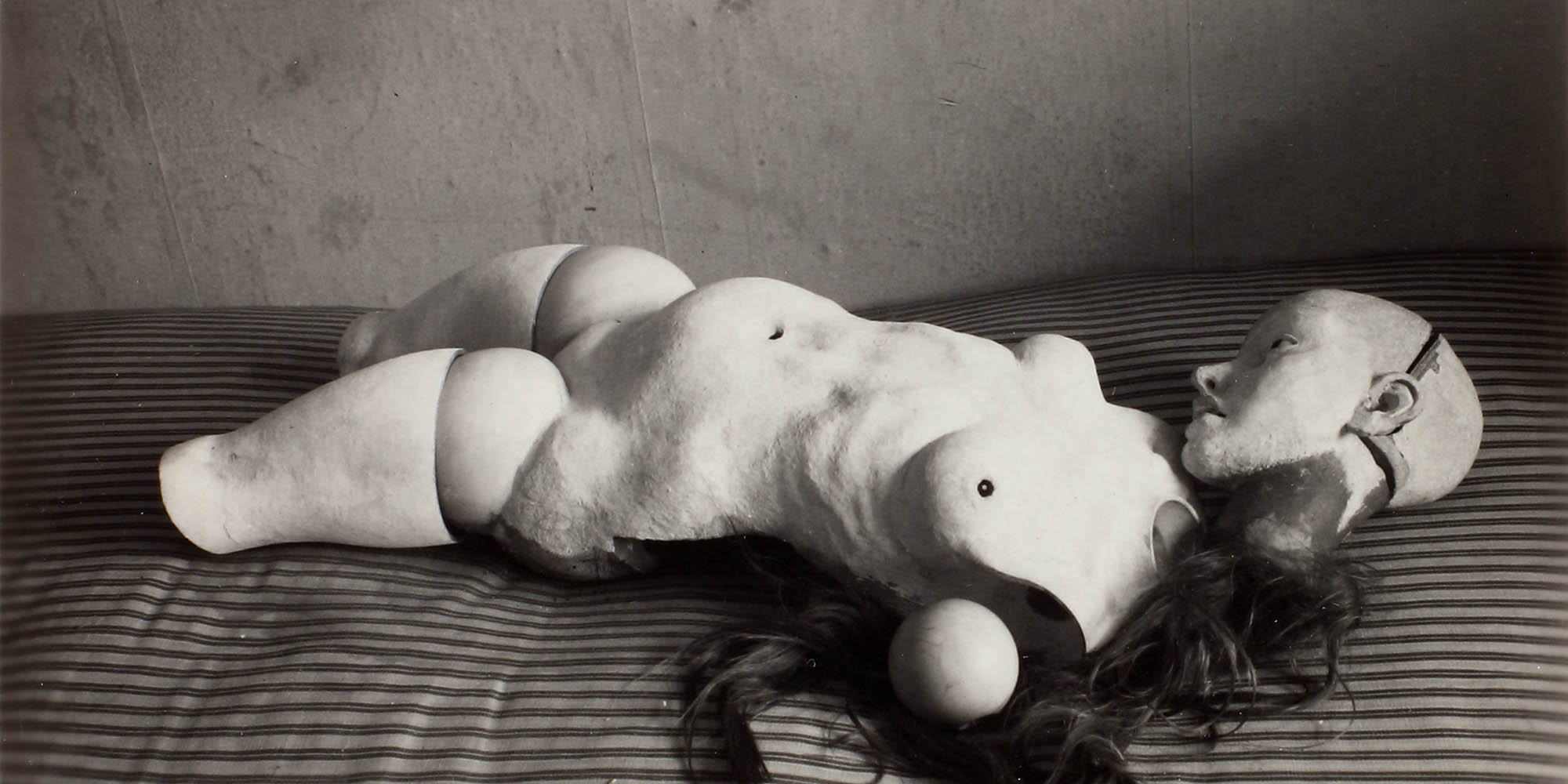 A photograph of a doll like figure laying on a striped surface, perhaps a bed. The digure has dark hair, and is nude with no arms of lower legs.