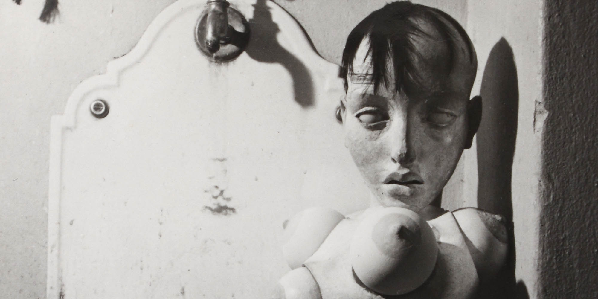 A banner showing a photograph of a sculpture or mannequin, which is a head with dark hair connect to a base with breast like objects attached to it. The doll is next to a wall, balanced on a sink with a spout at its top.