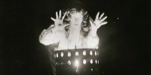 This is a work of art. Please note that as it is impossible to know the intent of the artist, the work is subject to interpretation. Each visitor may view and choose to understand the work differently. All effort has been made to provide a purely visual description. A black and white image of a woman with long wavy hair standing with her arms raised and her hands spread. She is standing over a chest high cylinder which has holes punched out, and it appears as if she is illuminated by something in the cylinder.