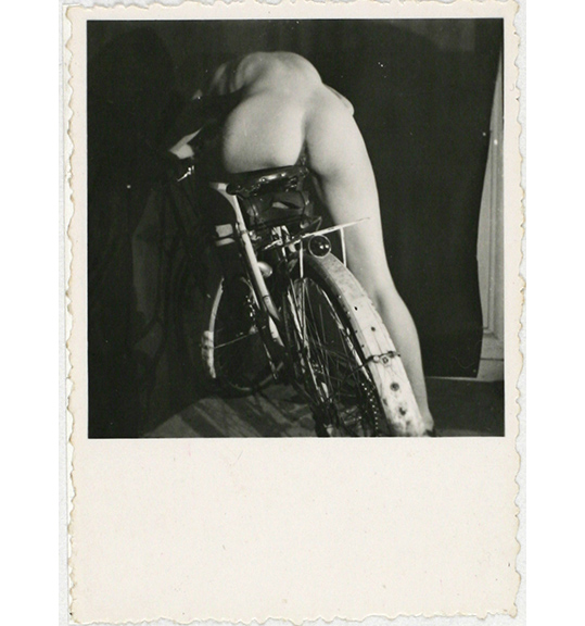This is a work of art. Please note that as it is impossible to know the intent of the artist, the work is subject to interpretation. Each visitor may view and choose to understand the work differently. All effort has been made to provide a purely visual description. A photograph of a woman seen from the back. The subject is nude and sits on the seat of a bicycle with the right leg on the ground. The photograph is on beige paper with a large margin at the bottom, and has torn edges.