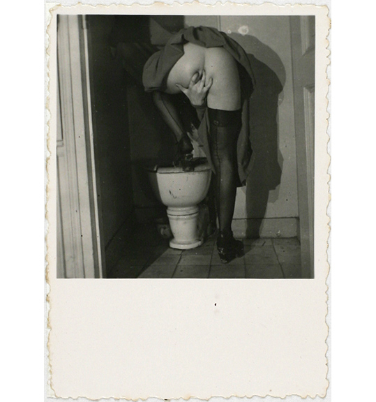 This is a work of art. Please note that as it is impossible to know the intent of the artist, the work is subject to interpretation. Each visitor may view and choose to understand the work differently. All effort has been made to provide a purely visual description. A black and white photograph of a figure standing with one leg up on a toilet, showing the nude backside. The figure wears black stockings and reaches the right hand between their legs, as if inserting a finger into their genitals. The photograph is on beige paper with a large margin at the bottom, and has torn edges.