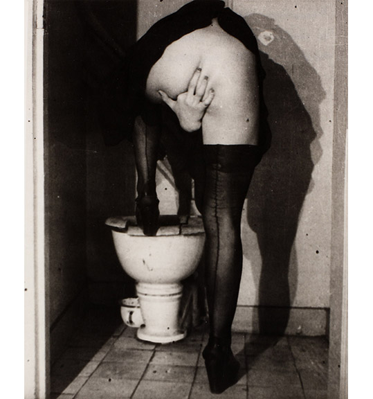 This is a work of art. Please note that as it is impossible to know the intent of the artist, the work is subject to interpretation. Each visitor may view and choose to understand the work differently. All effort has been made to provide a purely visual description. A black and white photograph of a figure standing with one leg up on a toilet, showing the nude backside. The figure wears black stockings and reaches the right hand between their legs, as if inserting a finger into their genitals.