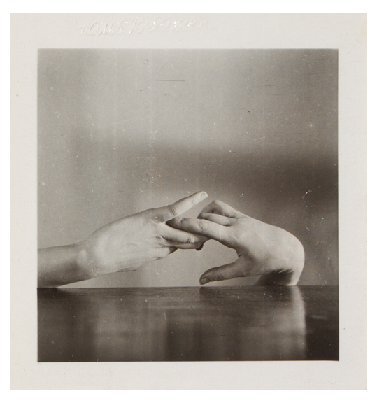 A photograph of two hands with ring and index fingers touching. The hand on the right is bent at the wrist and the hand on the left stretches to the right. Both hands appear in front of what appears to be a table.