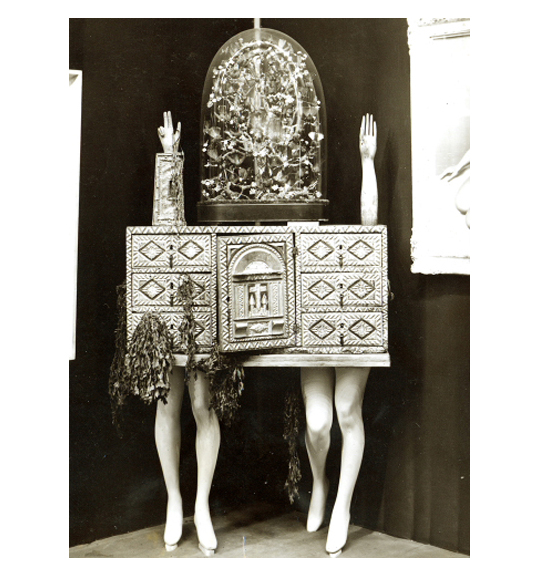 Image of a small chest, which rests on two pairs on mannequin legs, side by side. Dried plant matter hangs off of the chest’s drawers on the left side, and two mannequin arms are placed upright on the top of the chest, fingers pointing upwards.