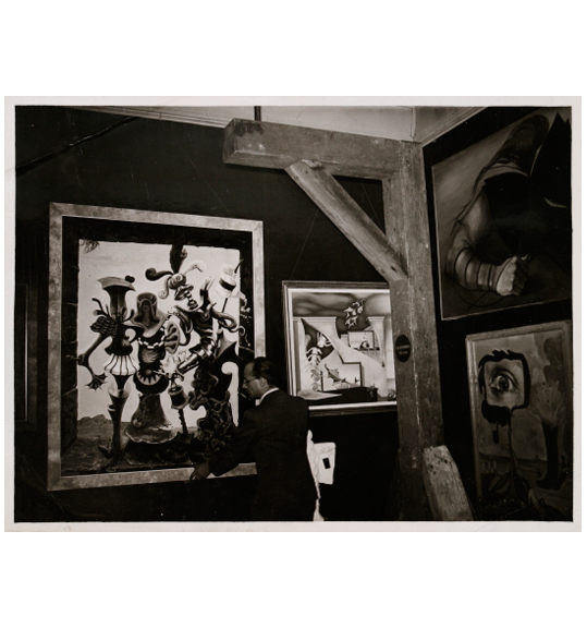 Photograph of a man in a dark suit standing next to what appears to be a wooden beam in the corner of a room. He stands in front of two artworks hanging on the wall. Two works of art hang vertically on the opposite wall.