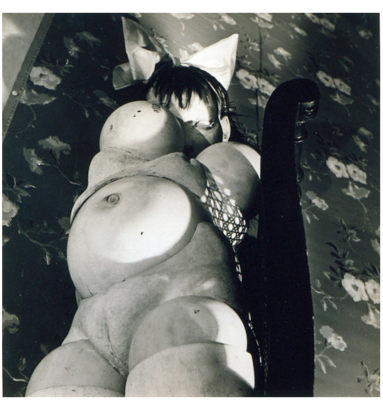 This is a work of art. Please note that as it is impossible to know the intent of the artist, the work is subject to interpretation. Each visitor may view and choose to understand the work differently. All effort has been made to provide a purely visual description. A black and white photo of a doll like mannequin. The angle shows the doll's stomach and chest, and one eye as well as a large bow on its head.