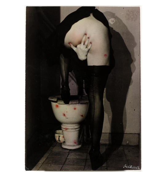 A photograph of a figure standing with one leg up on a toilet, showing the nude backside. The figure wears black stockings and reaches the right hand between their legs, as if inserting a finger into their genitals. There are several red dots on the image, mostly on the toilet.