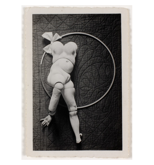 A photograph of a mannequin, which is lying on a large circle or hoop on top of a quilted fabric. The mannequin has ball joints and an arm and a leg. A bow is placed near the mannequin on the upper left side.