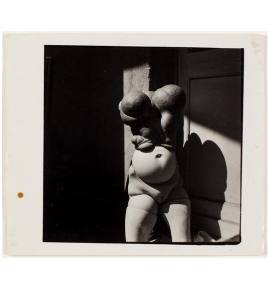 A photograph of a headless doll like figure with a nude body and segmented stomach, pelvis, and breasts. The doll's shadow is project on a wall on the right side.