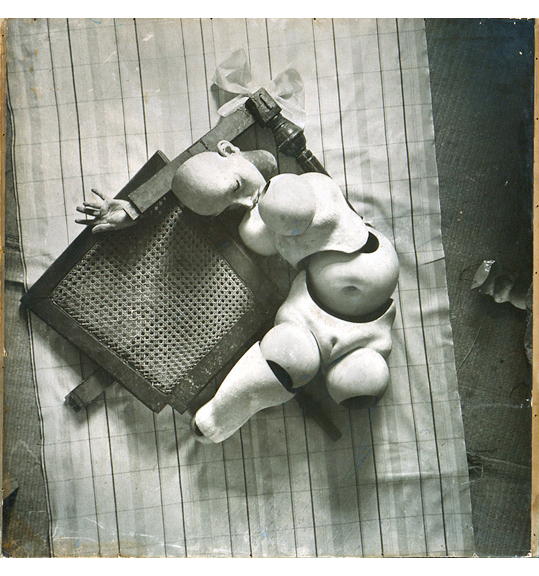 A photograph of a nude doll figure with segmented body parts. The body is curves to the right, has one leg that ends above the knee and one that ends at the thigh. The doll's arm appears under its head, and it lays on top of a checked fabric and what appears to be the woven seat of a wooden chair.