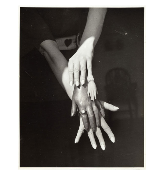 This is a work of art. Please note that as it is impossible to know the intent of the artist, the work is subject to interpretation. Each visitor may view and choose to understand the work differently. All effort has been made to provide a purely visual description. A photograph of what appear to be women's hands, stacked on top of each other. The bottom hand has white polished nails, and the hand on top of it wears a ring. The hand on top wears a small object on its pinky, which is a small hand itself.
