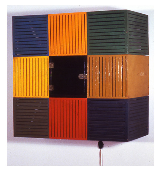 This is a work of art. Please note that as it is impossible to know the intent of the artist, the work is subject to interpretation. Each visitor may view and choose to understand the work differently. All effort has been made to provide a purely visual description. A photograph of a cube like object showing nine colorful sections in blue, orange, green, yellow, black, orange, brown, red, and purple. The colored sections appear to have long horizontal and vertical slits.