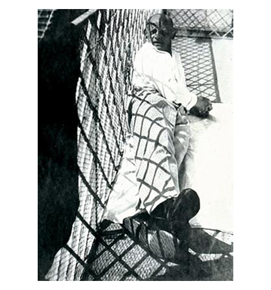 A black and white image of a man wearing light colored pants and shirt. The man is reclining by what appears to be a chain fence, which casts a shadow onto his body.