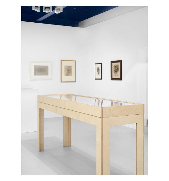 A photograph of an exhibition corner wall showing four framed works, two on the right wall and two on the left wall. A display case on four legs in a light colored wood stands in the center.