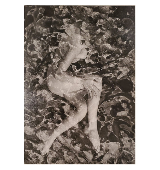 A photograph showing what appears to be a nude woman. The viewer is looking down, and she is sitting or lying down on her side, facing the right. Her legs are bent at the knee and she has her right arm resting below the thigh. Her face is hidden by her hair and there is an overall watercolor or marble effect.