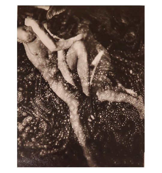 A photograph of a nude couple. The man sits with one leg outstretched and the other bent, and the woman embraces his neck as she lies on his left side.